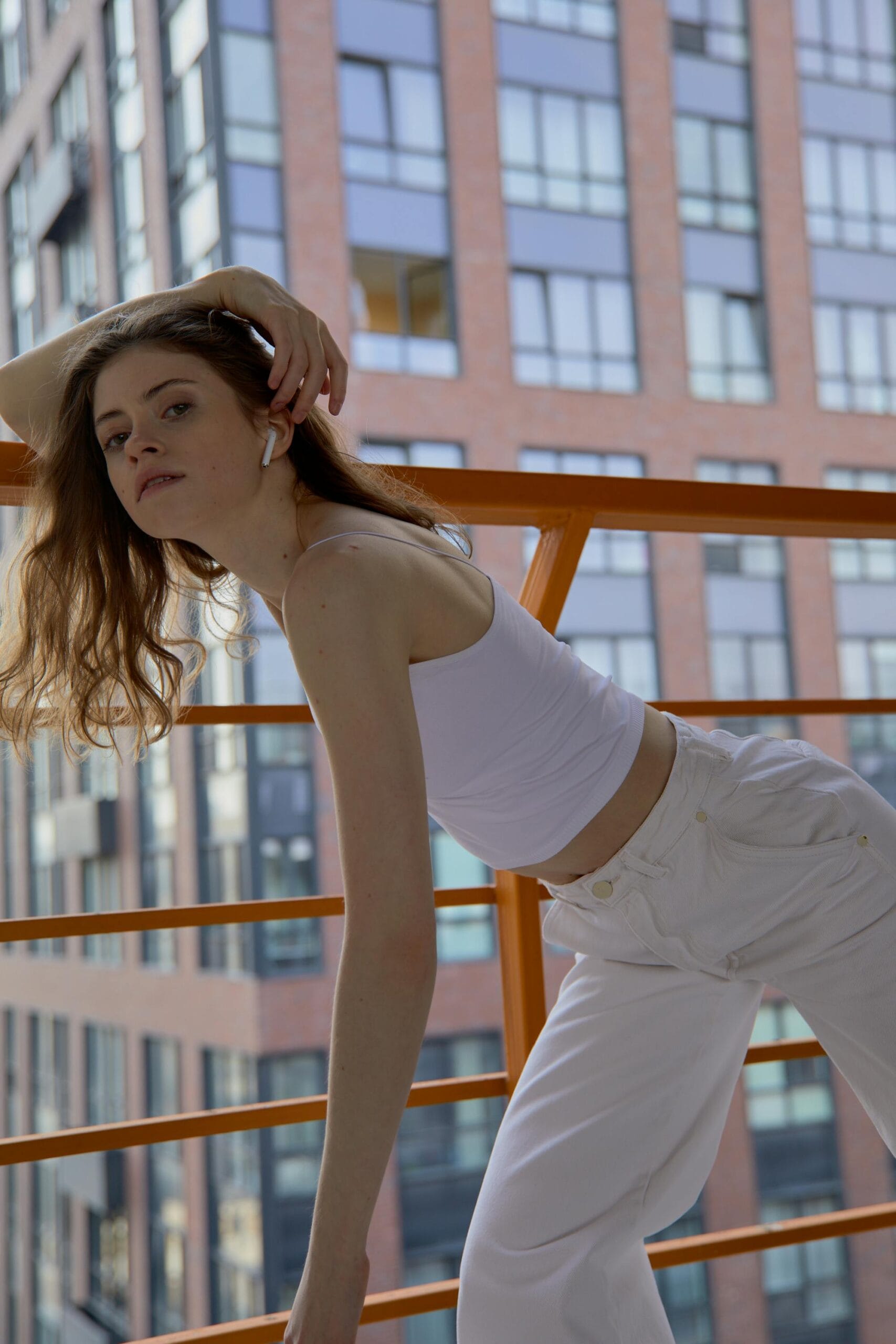 Stylish woman in a white outfit posing with Airpods on a city balcony, showcasing modern urban fashion.