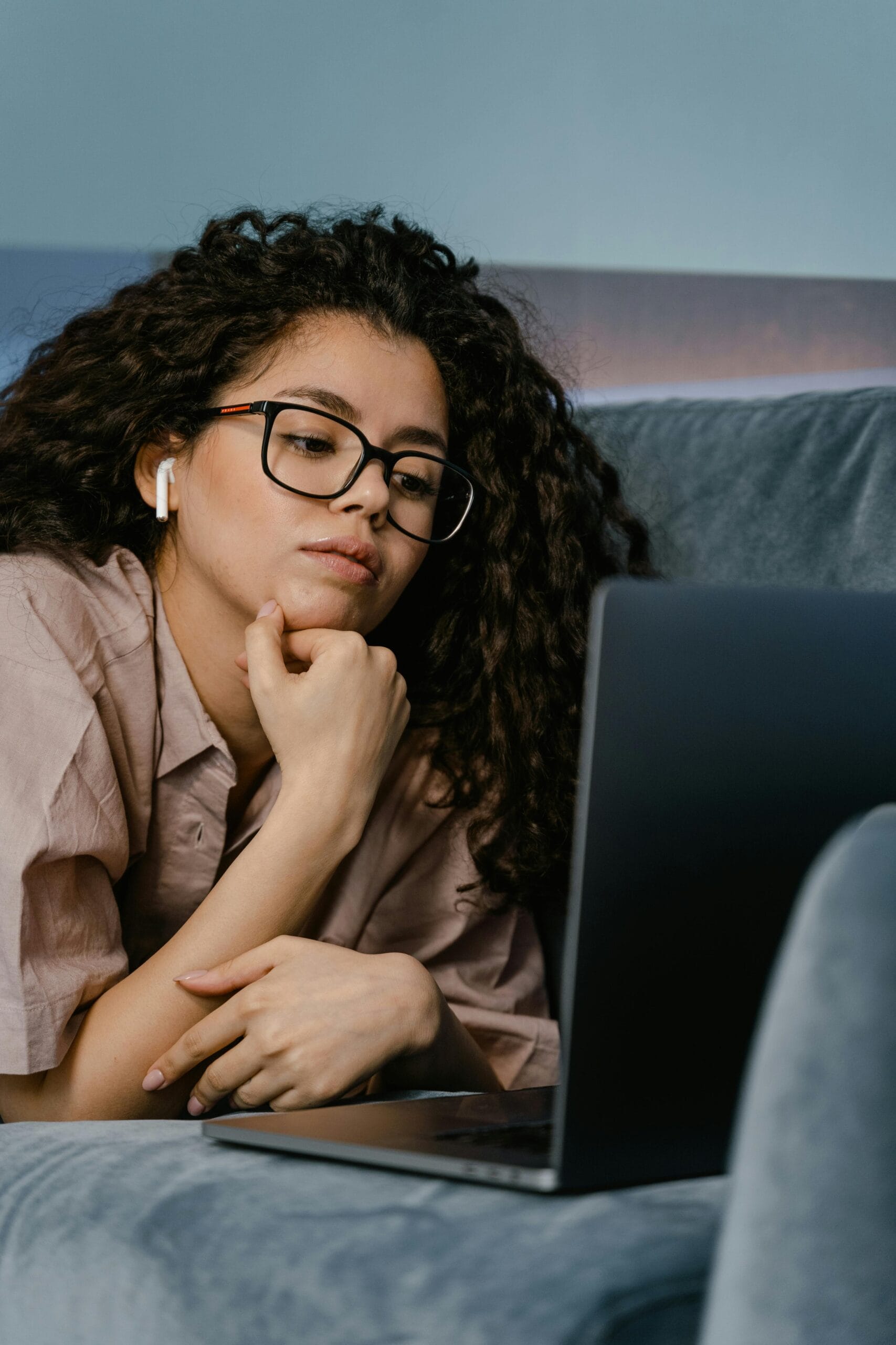 A woman in glasses working remotely on a laptop while lying on a couch at home.
